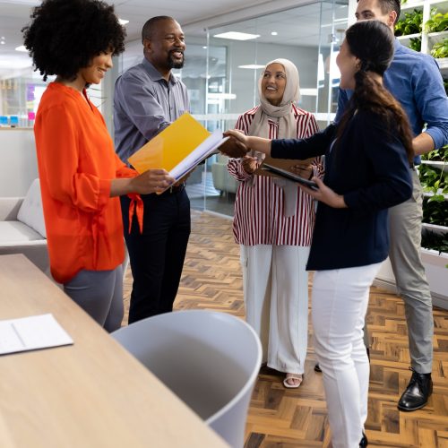 Vertical image of happy diverse female and male businesspeople talking and shaking hands in office